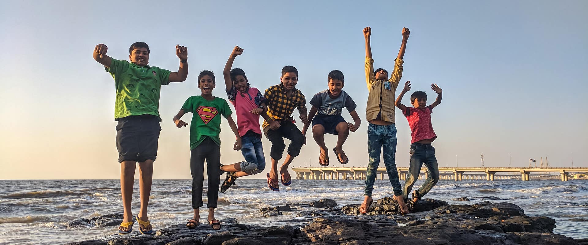 Children jumping on rocks by the sea at sunset.
