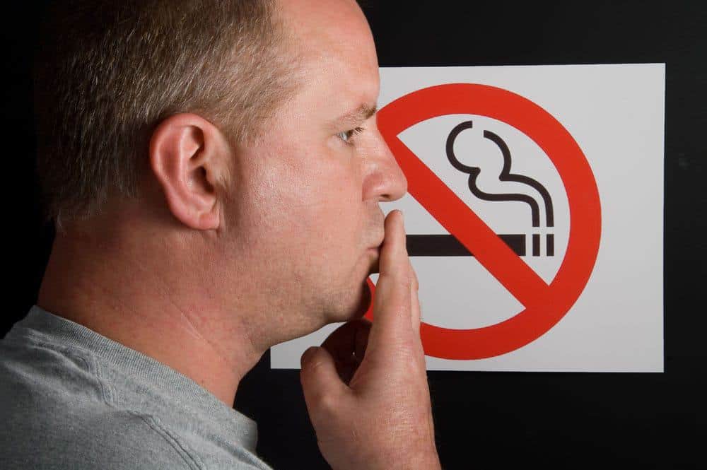 Man standing beside a no smoking sign with hand near mouth.